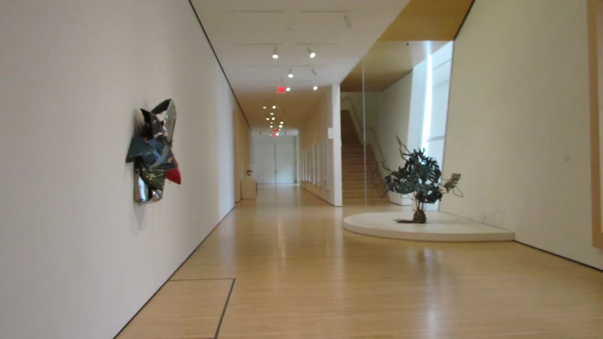 A long, minimalist hallway in a modern art museum with white walls, light wood floors, and sparse abstract sculptures. On…