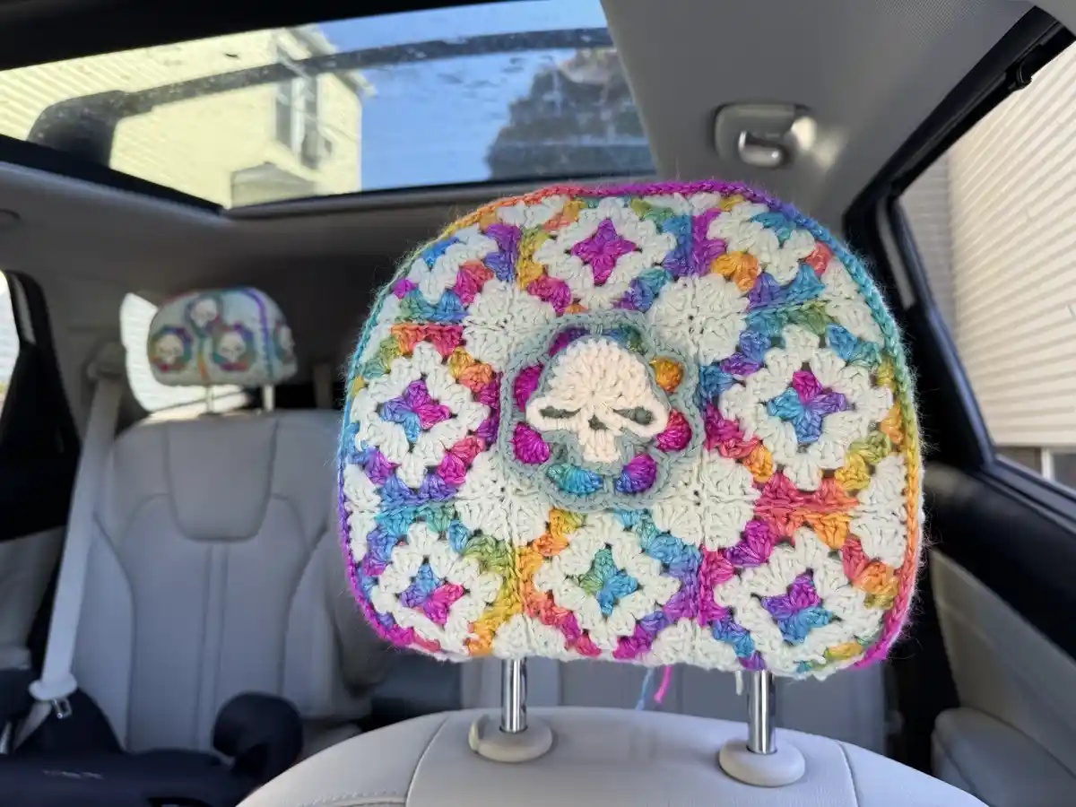 A close-up photo shows a colorful, hand-crocheted headrest cover on a car seat, featuring a central skull motif surrounded…