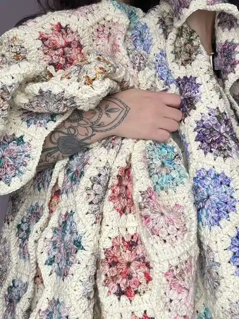 A person is wrapped in a colorful, hand-crocheted blanket made of floral granny squares in pastel shades of pink, purple…