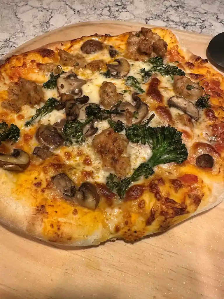 A close-up photo of a homemade pizza on a wooden board, featuring melted cheese, sausage pieces, sliced mushrooms, and…