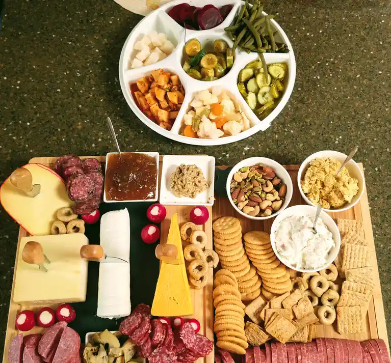 An overhead shot of an elaborate charcuterie board with multiple cheeses, meats, crackers, nuts, dips, and a round…