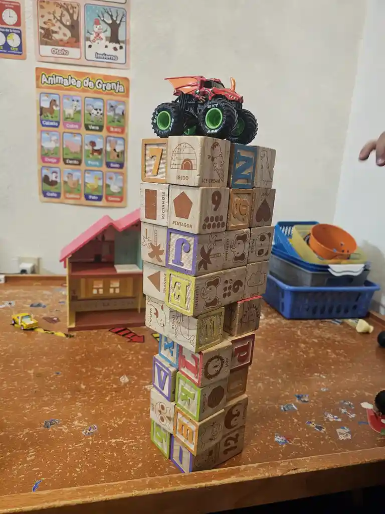 A red monster truck toy with green wheels is perched atop a tall, precarious tower of wooden blocks featuring letters…