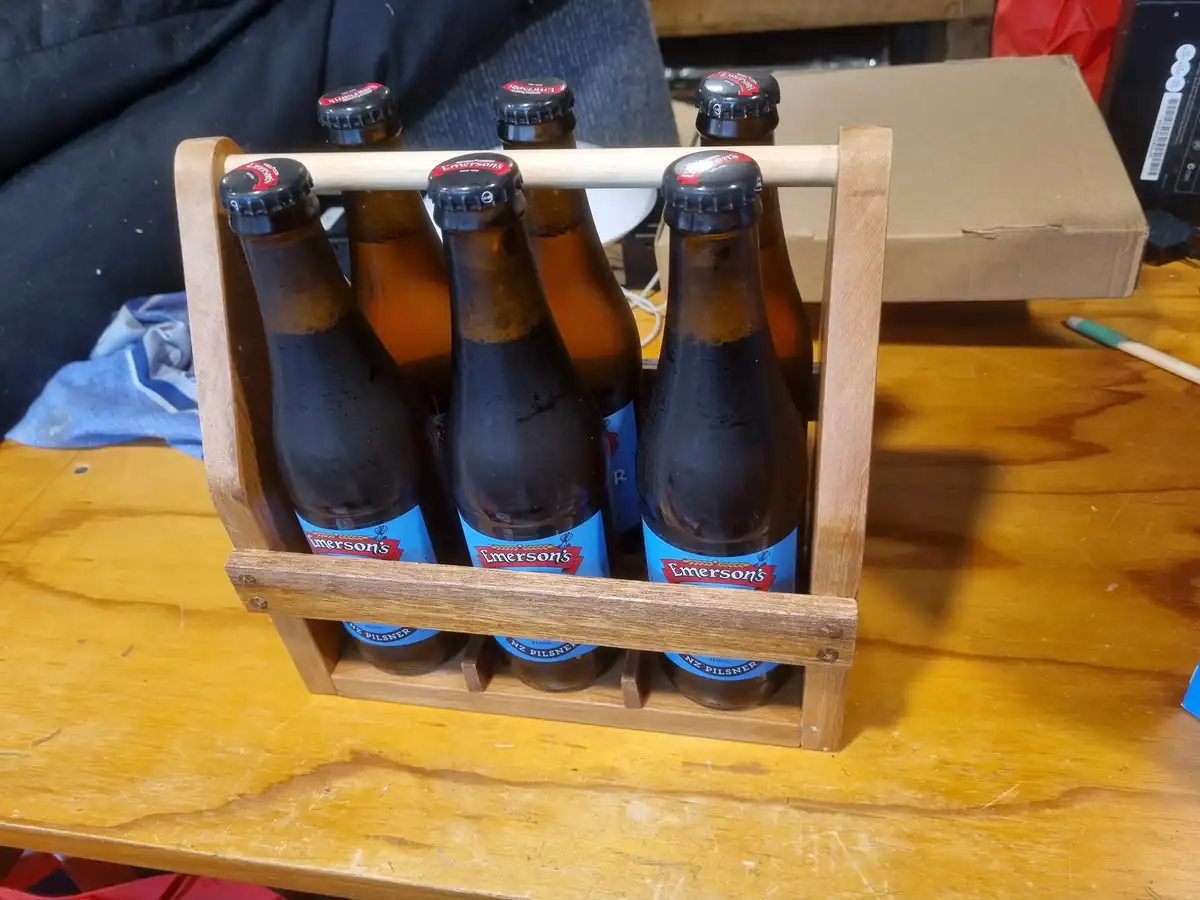 A wooden beer carrier holds six brown glass bottles of Emerson's NZ Pilsner, each with a black cap and blue label, sitting…