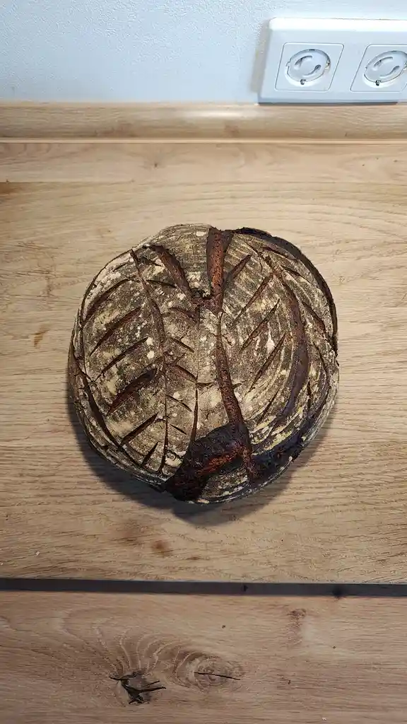 A round loaf of bread sits on a wooden countertop, its crust deeply charred and cracked in a pattern resembling tree bark or…