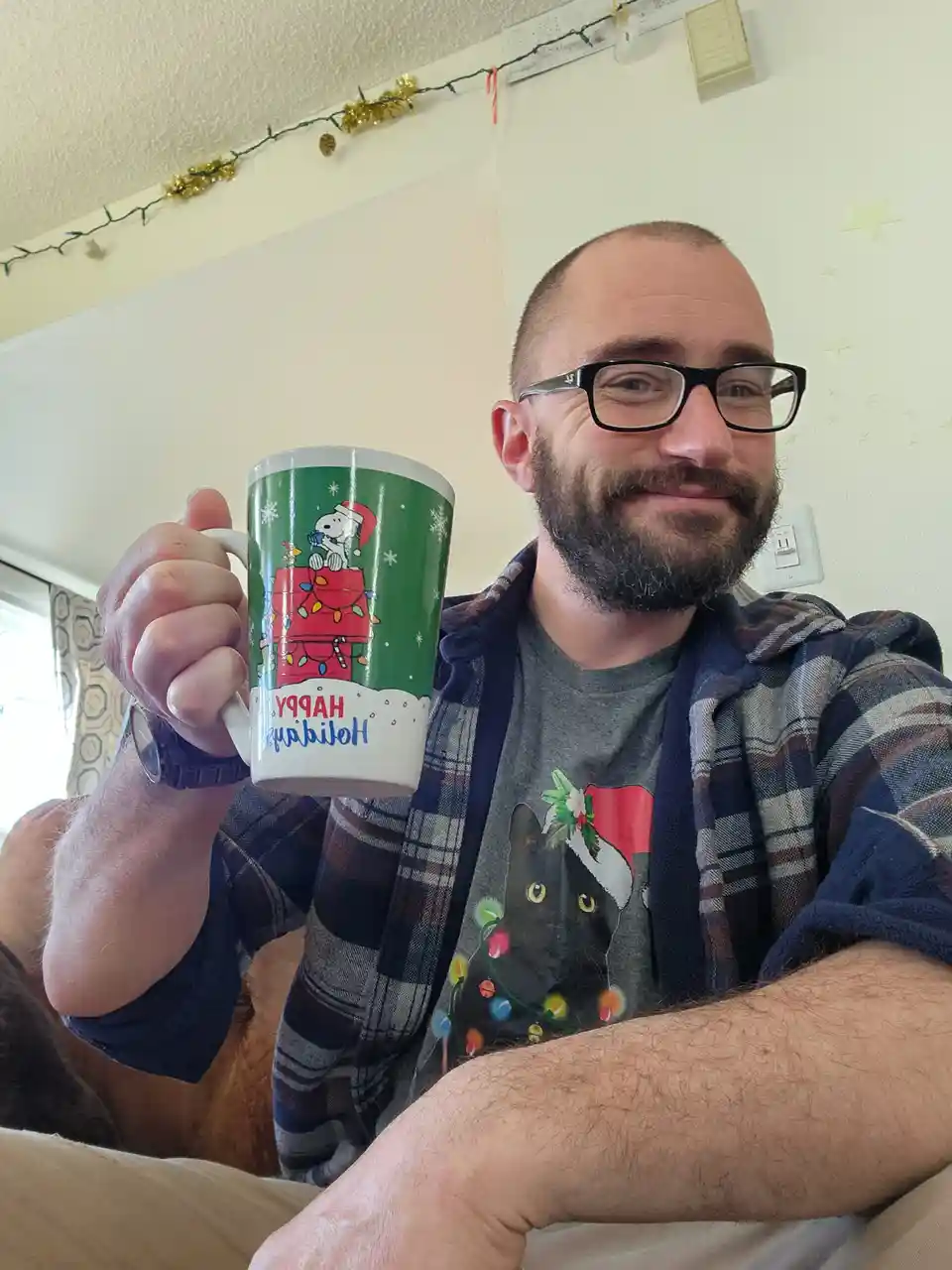 A man with glasses and a beard holds up a green holiday mug featuring Snoopy on a decorated doghouse with the text 'Happy…