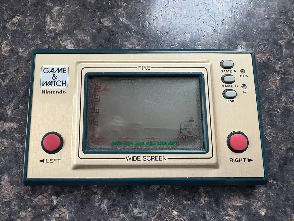 A vintage Nintendo Game & Watch handheld console with a dirty screen showing a fire scene and an ambulance. The device has…