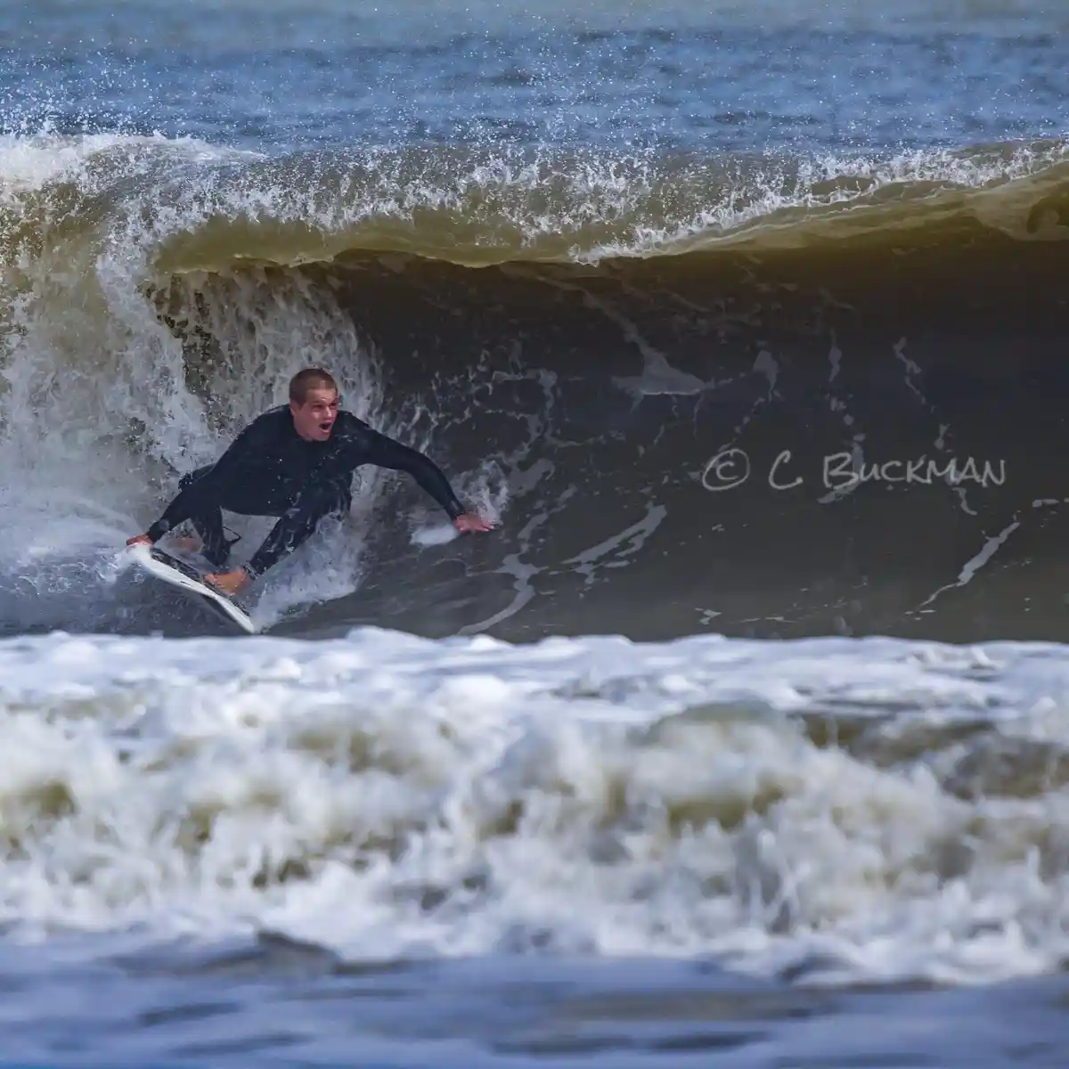 A surfer in a black wetsuit rides inside a curling ocean wave, looking shocked or scared with his mouth open. The wave is…
