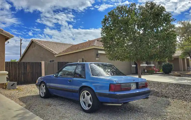 A blue 1990s Ford Mustang LX is parked on gravel in front of a suburban house with a large tree and a blue sky with clouds…