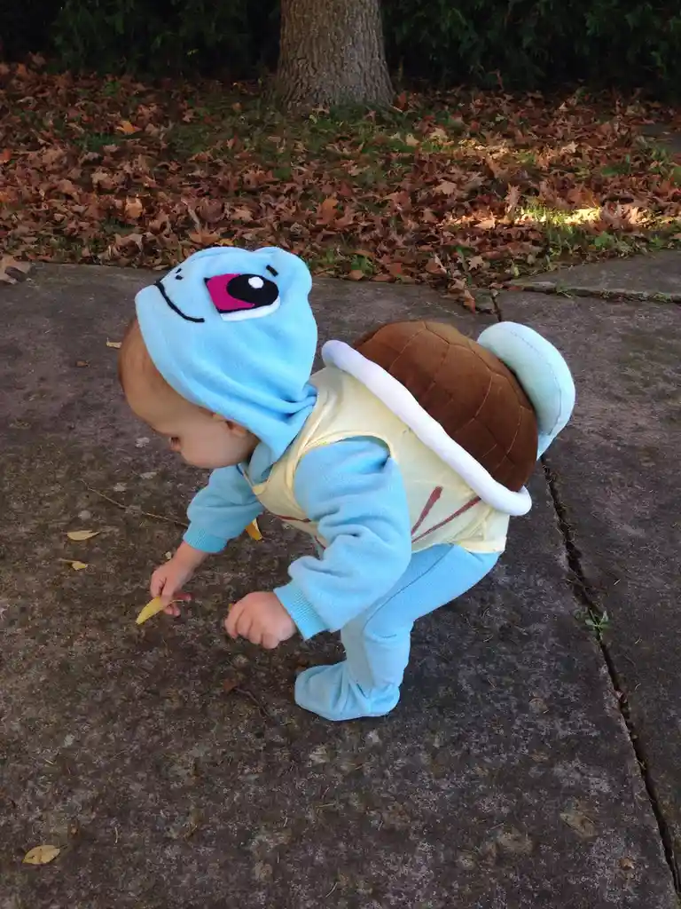 A baby wearing a full-body Squirtle costume, including a hood with a large cartoon face and a brown shell attached to the…