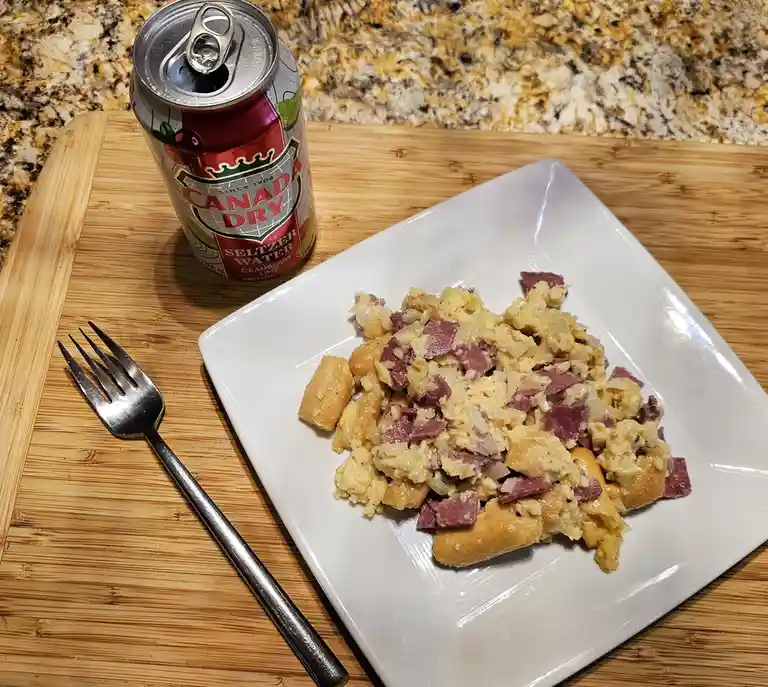 A white square plate holds a dish of scrambled eggs mixed with gnocchi and chopped corned beef, sitting on a wooden cutting…