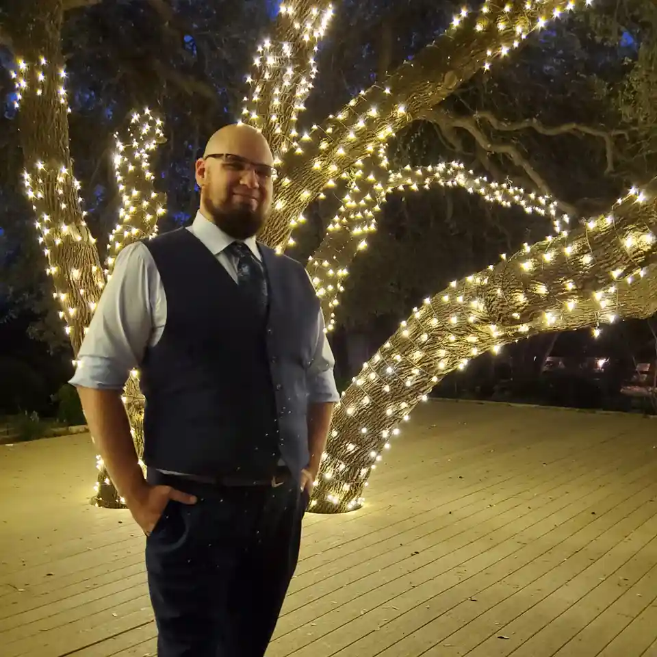 A bald man with a beard and glasses, wearing a vest, shirt, and tie, stands confidently with hands in pockets on a wooden…
