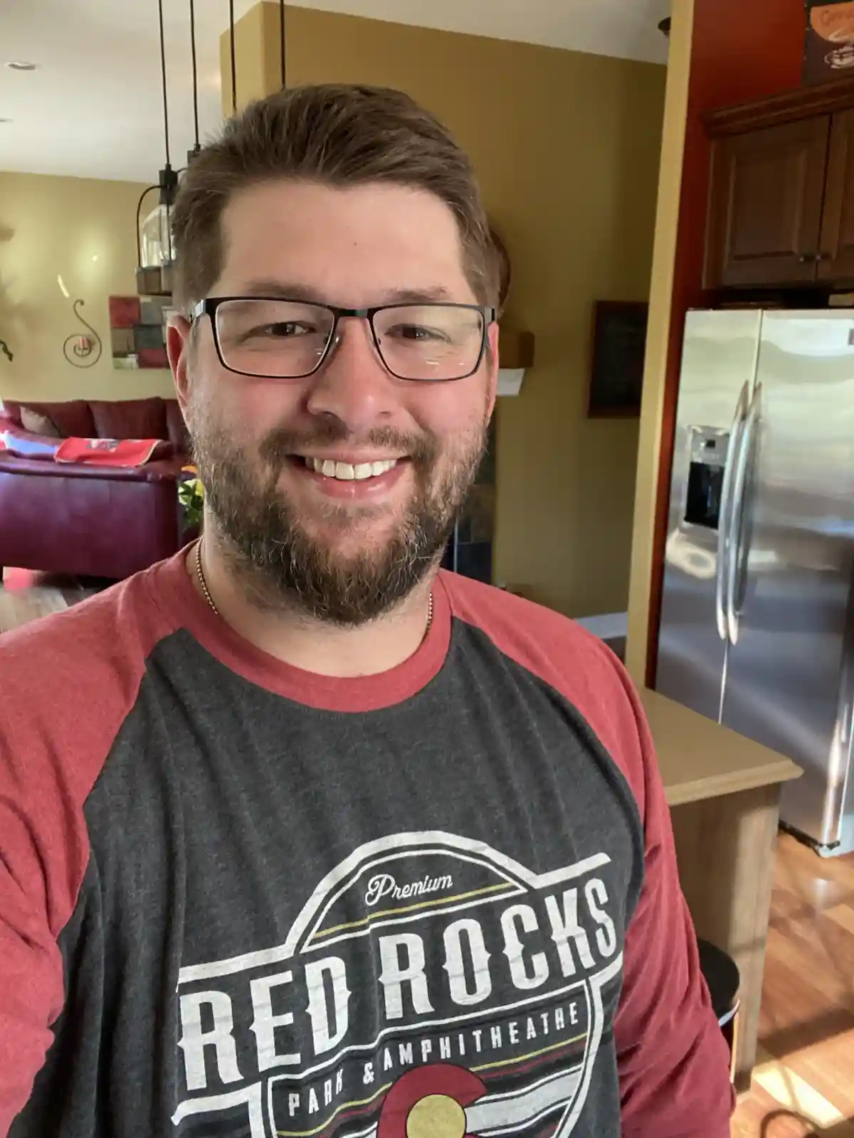A smiling man with glasses and a beard wears a red and grey t-shirt featuring the 'Red Rocks Park & Amphitheatre' logo. The…