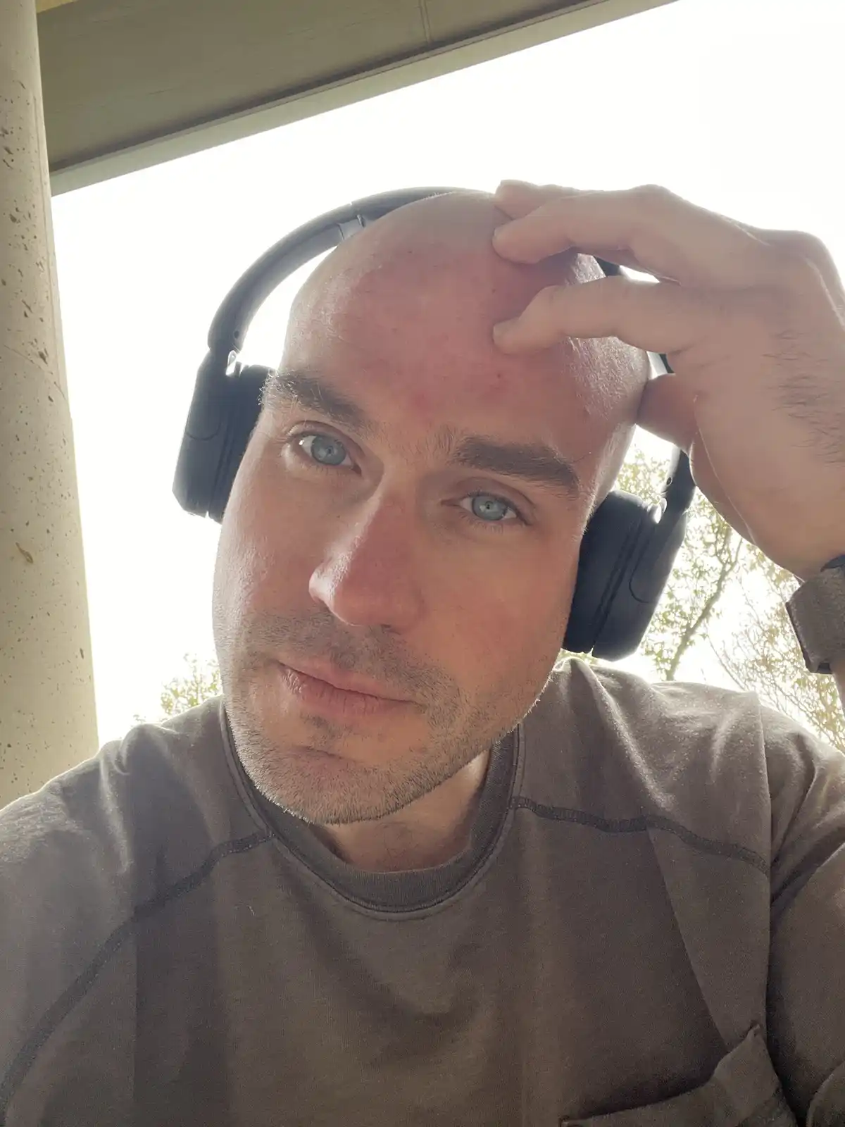 A bald man wearing black over-ear headphones looks directly at the camera with a neutral expression while his right hand…