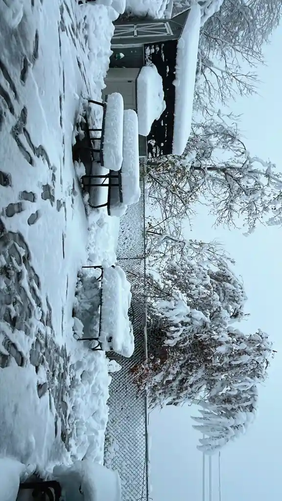 A snowy outdoor scene with a picnic table and benches completely buried under thick snow, footprints visible in the snow…