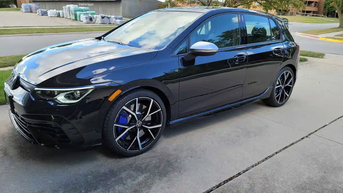 a glossy black volkswagen golf r is parked on a concrete driveway, featuring sporty multi-spoke wheels with blue brake…