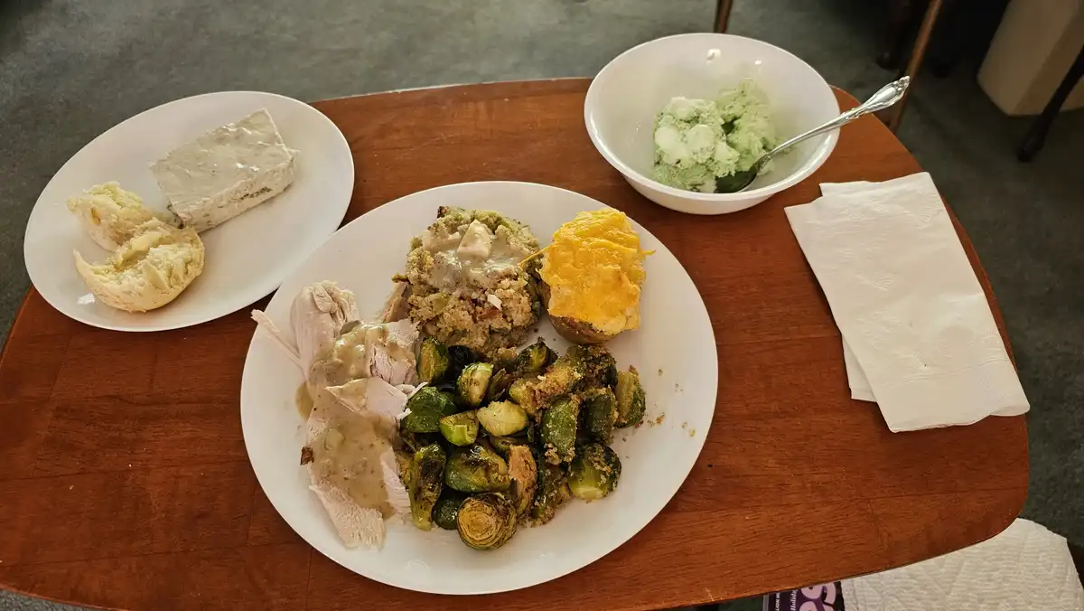 A wooden table holds a large plate of Thanksgiving food including turkey with gravy, stuffing, roasted Brussels sprouts, and…