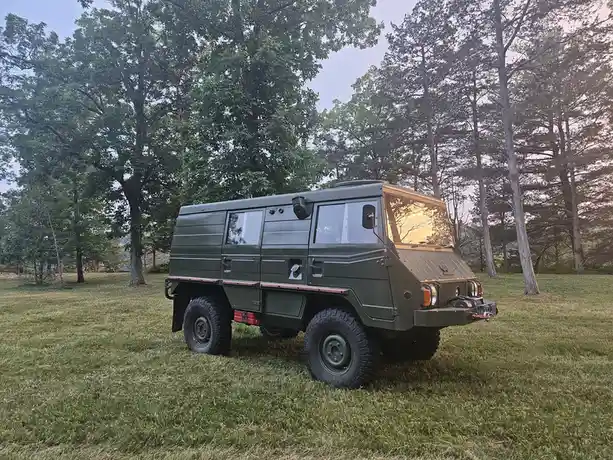 A green, boxy, military-style vehicle with large tires and a utilitarian design is parked on a grassy lawn surrounded by…