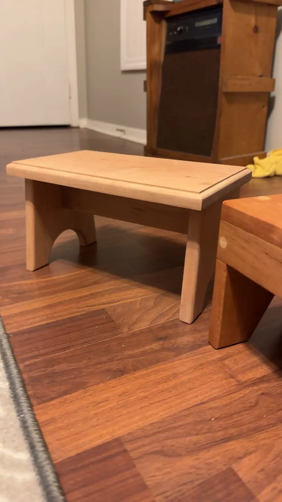 A simple, light-colored wooden footstool with arched legs sits on a hardwood floor next to a darker wooden piece of…