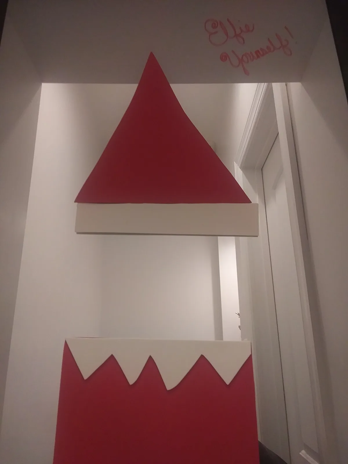 A doorway is decorated with a large, red, triangular Santa hat cutout with a white brim, and a red, jagged-edged lower…