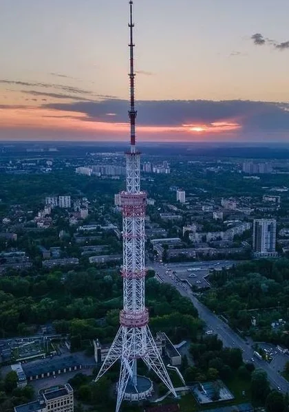 An aerial photo of a tall, white lattice tower with red accents, standing in a cityscape at sunset. The sky is orange and…