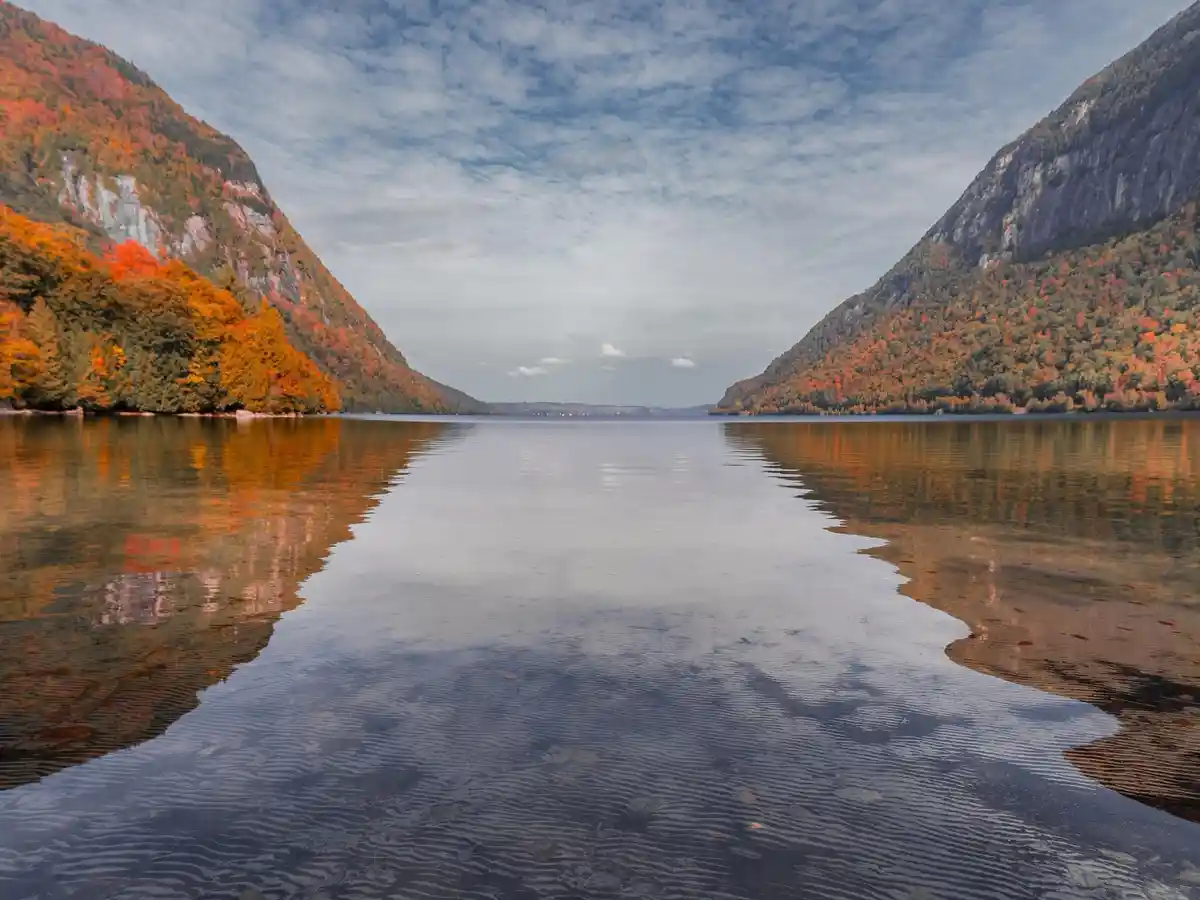 A serene autumn landscape featuring a calm lake flanked by steep mountains covered in vibrant orange and red foliage, with…