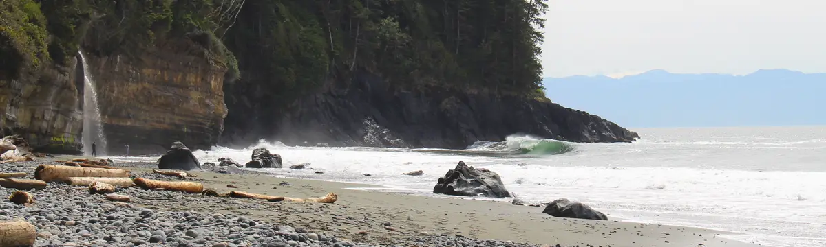 A scenic coastal beach with a small waterfall cascading down a cliff into the ocean, waves crashing nearby, and driftwood…