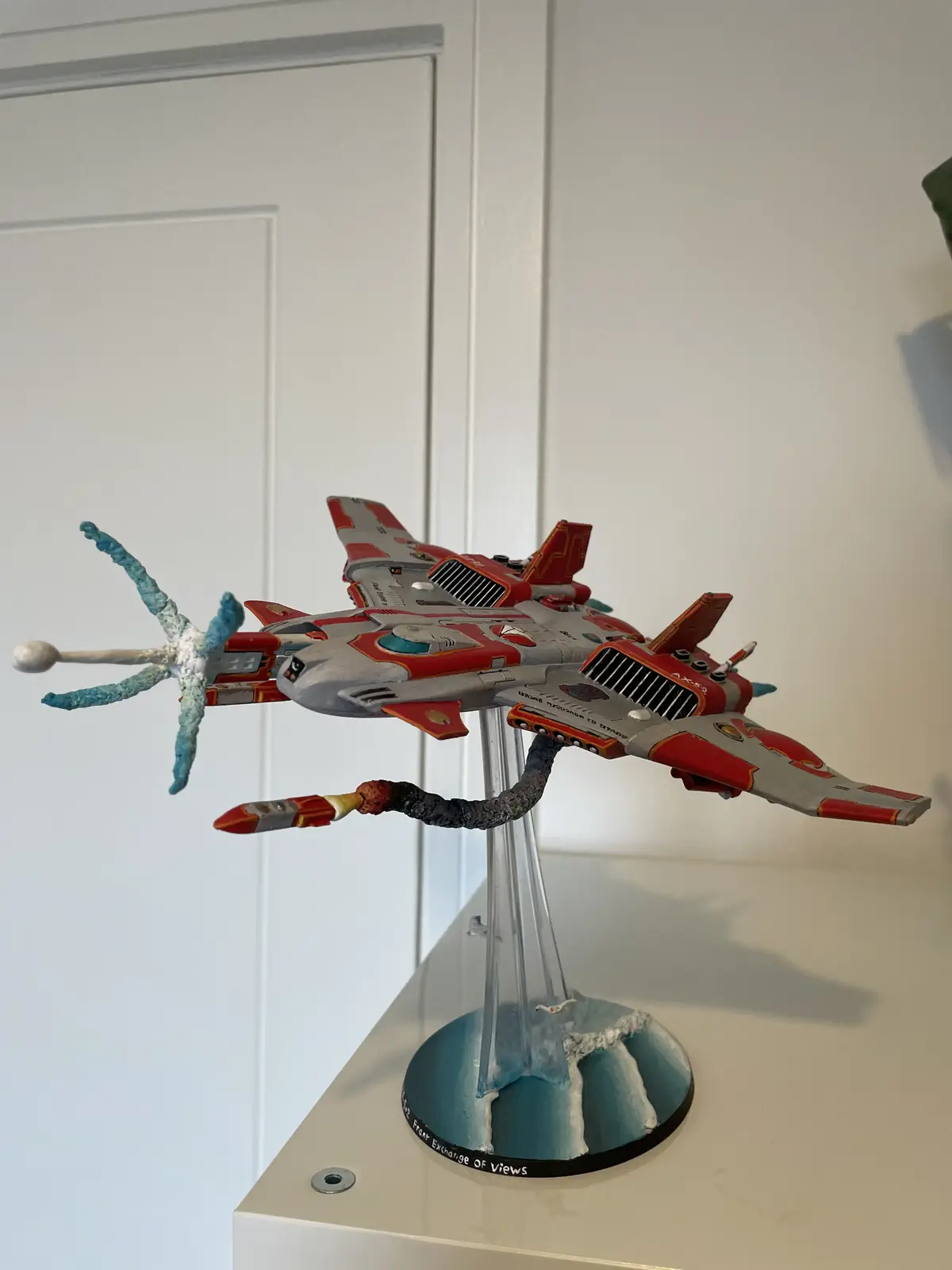A red and silver spaceship model is mounted on a clear stand with a blue and white painted base, positioned on a white shelf…