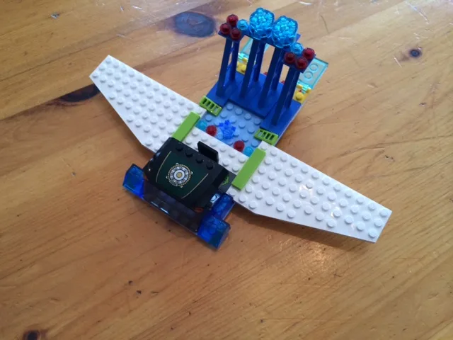 A LEGO model of an airplane with a white body, blue engine, and blue and red propellers sits on a wooden table. The cockpit…